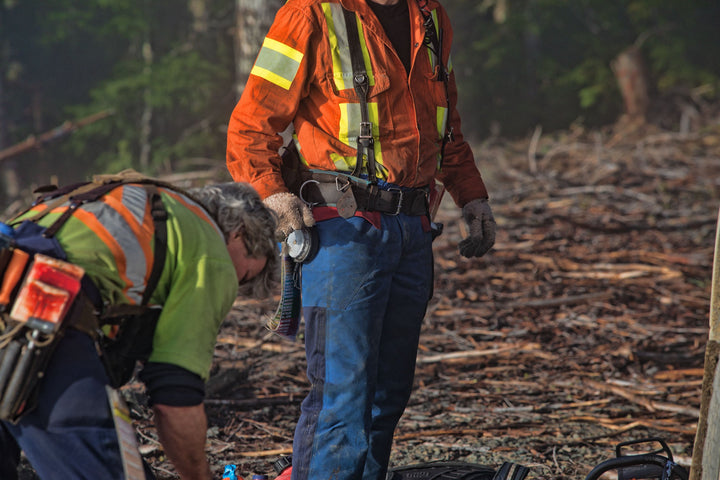 Two fallers working on a cut block, one standing in blue saw pants with wedges and a tape on his belt, the other kneeling over gear