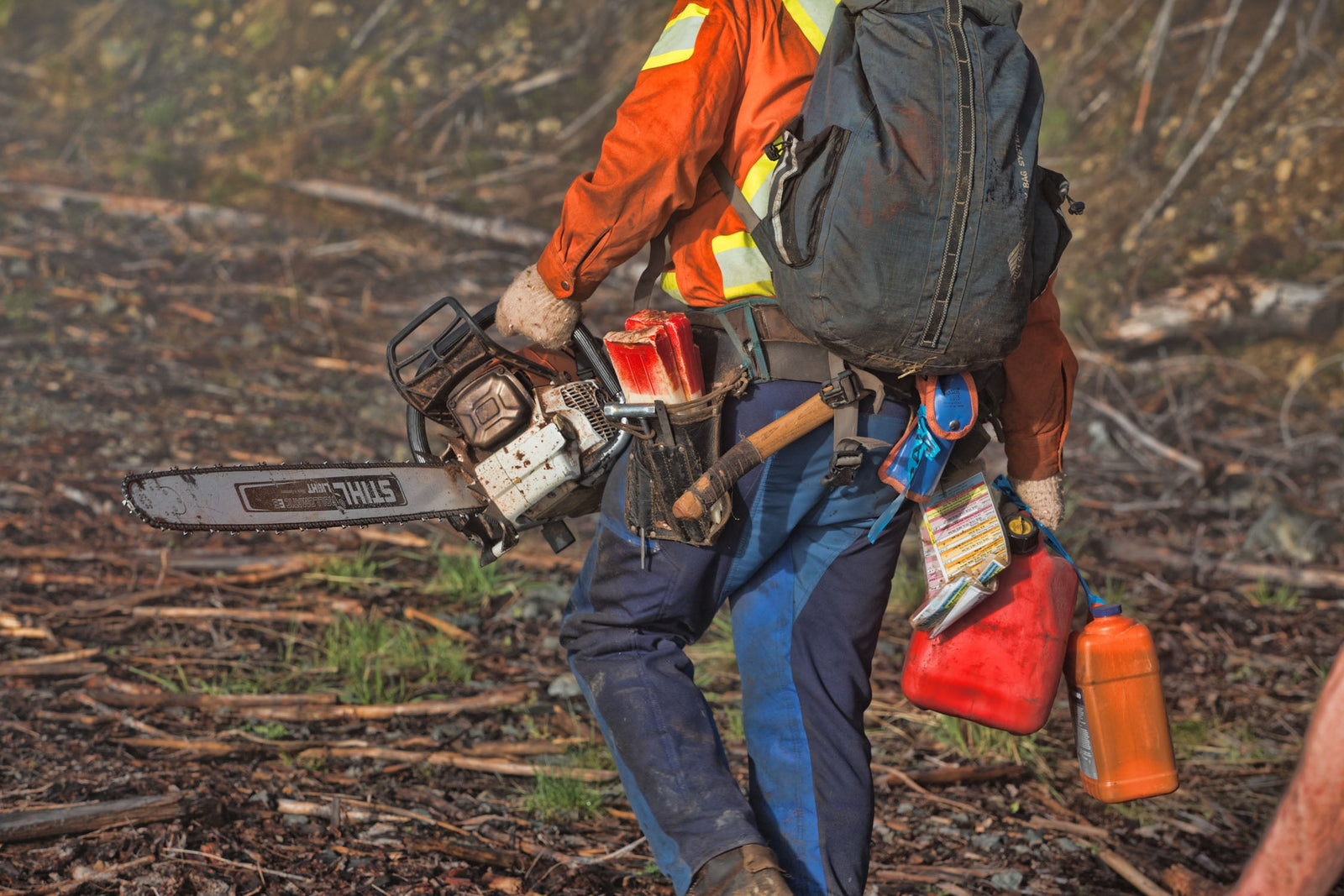 Faller walking out of the cut carrying his chainsaw, wedges and fuel cans, wearing blue saw pants